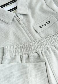 Vaaleanharmaa haalari, jossa on teksturoitu kuvio, vetoketjullinen etuosa, kaulus, joustava vyötärönauha ja merkintätarra, jossa lukee "BAKER".