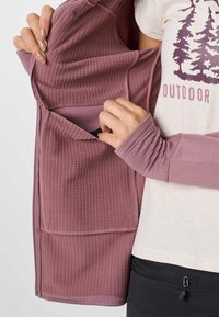 Hände zeigen die Innentasche einer mauvefarbenen, gerippten Jacke, getragen über einem weißen "Outdoor"-T-Shirt und schwarzen Hosen mit Reißverschlusstasche.