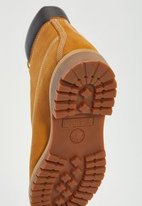 Suède gele laars met een zwarte rubberen neuskap. De buitenzool heeft een robuust patroon en het Timberland-logo. Zichtbare stiksels in lichte kleur.