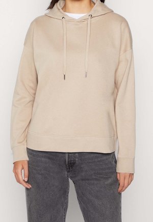 Beige hættetrøje med snore, båret over en hvid skjorte, kombineret med slidte sorte jeans.