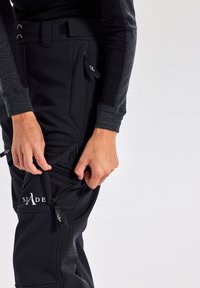 Zwarte sportieve broek met een ritszak, gemaakt van rekbaar materiaal. Heeft een taps toelopend ontwerp en een logo op de dij.