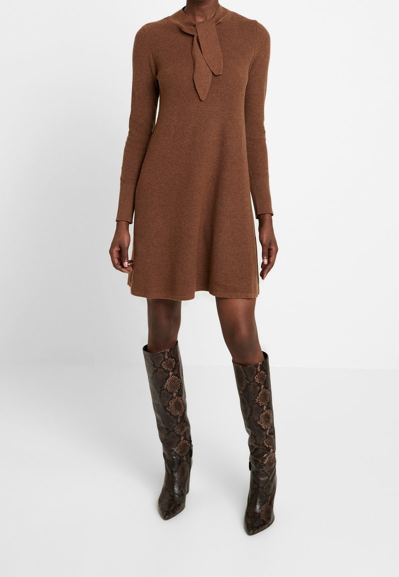 Femme portant une robe en maille marron à manches longues avec un détail de nœud au col, associée à des bottes montant jusqu'au genou à motif peau de serpent.