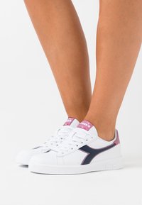 Vita sneakers med en strukturerad yta, marinblå sidologotyp och rosa accenter på hälen och tungan; med platta snörning och gummisula.