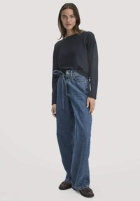 Navyblaues Langarmshirt kombiniert mit lässigen, hochtaillierten blauen Jeans aus Denim mit einem Gurt-Detail. Das Model trägt braune Schuhe und steht vor einem neutralen Hintergrund.