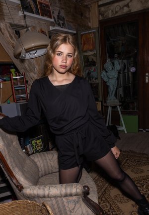 Zwarte wikkeloutfit met lange mouwen en shorts, met een geaccentueerde taille. Gevestigd in een rustieke kamer met vintage decor en een beeldhouwwerk.
