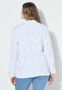 Frau mit welligem Haar, die ein strukturiertes weißes Langarmshirt und eine hellblaue Jeans trägt, steht vor einem schlichten weißen Hintergrund und ist von hinten zu sehen.