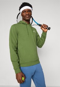 Grønn bomullshoodie med lomme foran og snørehette, parret med lys blå bukser. Tennisracket i hånden, holder en tennisball.