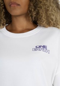 White cotton t-shirt featuring a purple "ONE EARTH" logo on the left side. Crew neckline with soft texture, short sleeves.