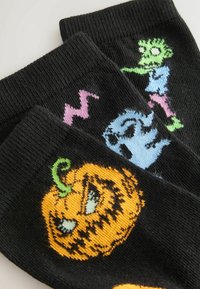 Strikkede sorte sokker med farverige Halloween-motiver: et grønt monster, et blåt spøgelse og et grinende orange græskar med grønne accenter.