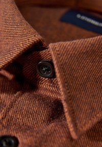 Zbliżenie na koszulę w kolorze rdzy z wzorem jodełki, czarnym guzikiem i miękką, teksturowaną tkaniną oraz kołnierzykiem z wierzchołkiem.