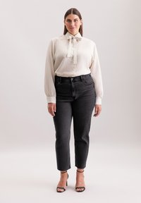 Blusa a coste color beige chiaro con dettaglio al collo a fiocco, abbinata a jeans grigio scuro a vita alta e sandali neri con tacco.
