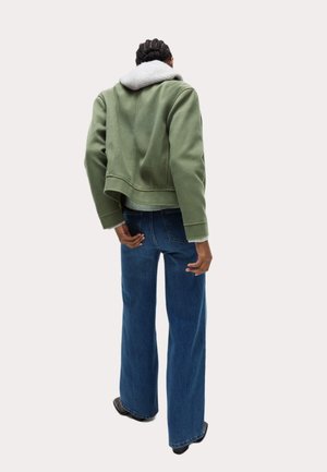Giacca verde corta con cappuccio grigio, abbinata a jeans a gamba larga blu. La giacca presenta un tessuto testurizzato e orlo aperto.
