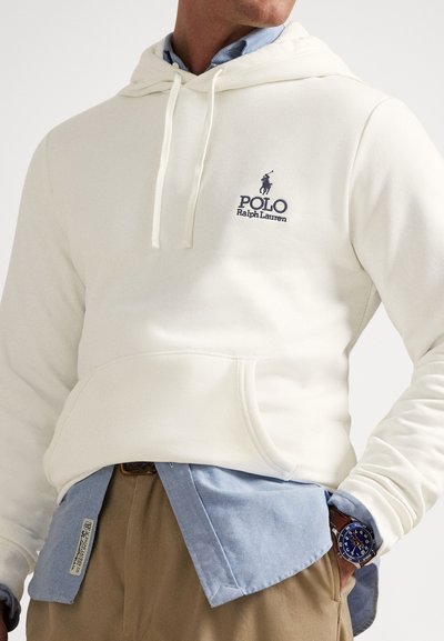 Λευκό φούτερ με κουκούλα και τσέπη καγκουρό, με το λογότυπο "Polo Ralph Lauren" σε σκούρο μπλε. Συνδυάζεται με ανοιχτό μπλε πουκάμισο από κάτω.