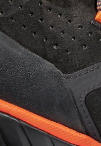 Scarpa sportiva nera con una combinazione di superfici lisce e testurizzate, con accenti arancioni lungo la suola e dettagli sulla parte superiore.
