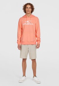 Kapuzenpullover in Koralle, mit einer vorderen Kängurutasche und weißem O'Neill-Logo. Kombiniert mit beigen Shorts und weißen Sneakers.
