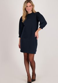 Marineblauwe sweatshirtjurk met een losse pasvorm, opgeblazen mouwen en een subtiel logo op de borst. Gladde stof met geribbelde boorden en zoom.