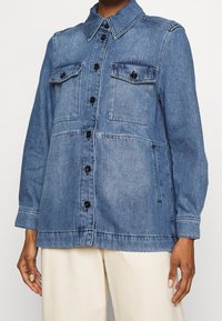 Chaqueta de mezclilla en azul medio; cuenta con cierre de botones, dos bolsillos en el pecho y mangas largas con puños. Tejido texturizado, diseño casual.