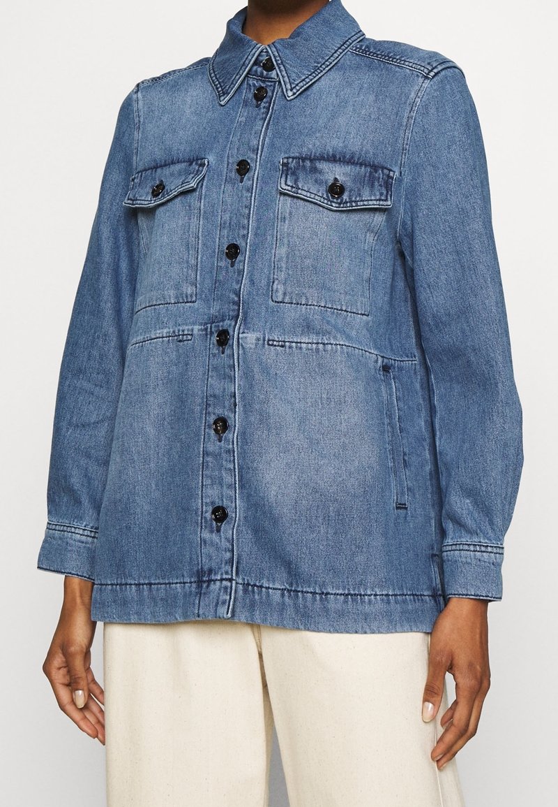 Chaqueta de mezclilla en azul medio; cuenta con cierre de botones, dos bolsillos en el pecho y mangas largas con puños. Tejido texturizado, diseño casual.