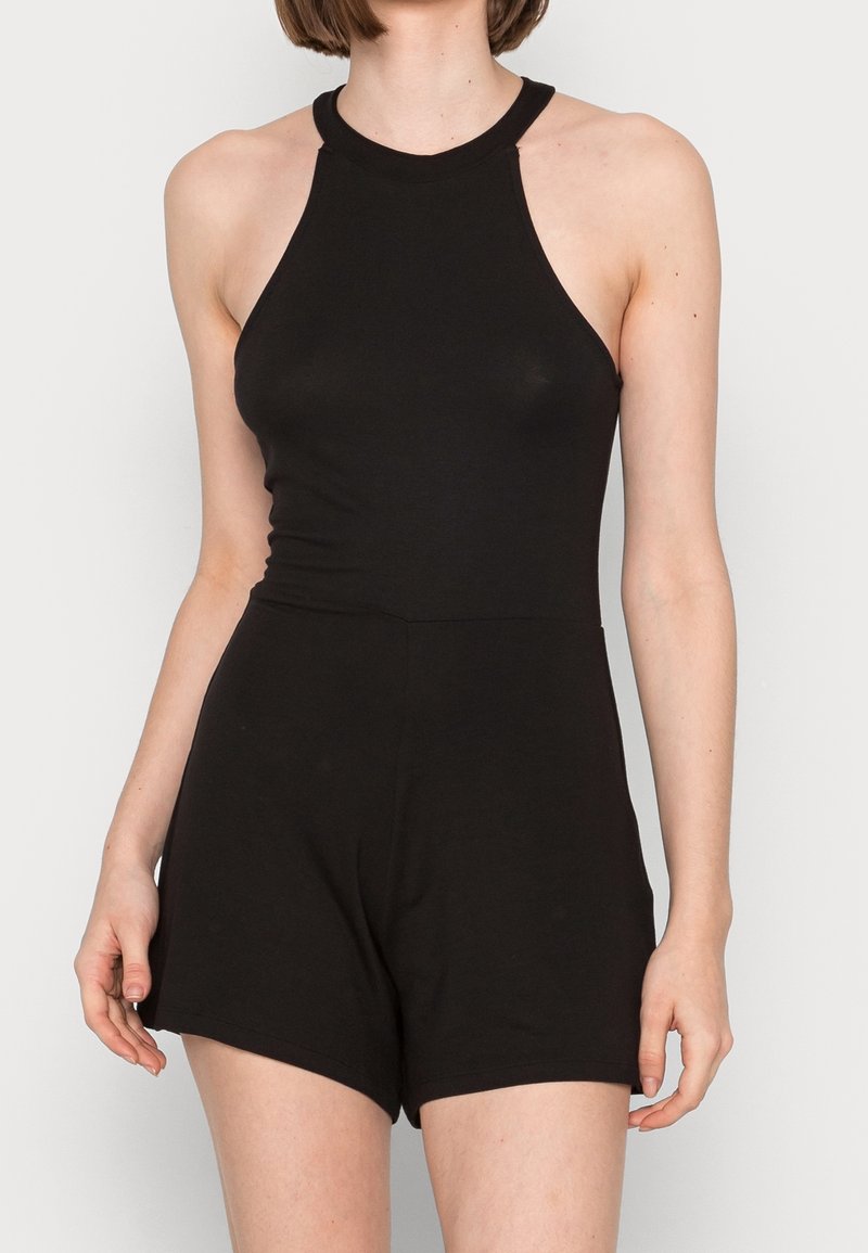Mujer vestida con un mono negro sin mangas y con cuello halter, que tiene la cintura ajustada y pantalones cortos, contra un fondo blanco liso.