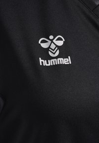 Schwarzer Stoff mit weißem, stilisiertem Bienen-Logo und dem Schriftzug „hummel“ auf dem Brustbereich eines Kleidungsstücks gedruckt.