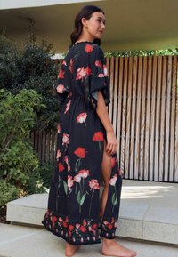 Een vrouw staat op stenen trappen in een lange zwarte bloemenrobe, waarbij ze een been laat zien door een split, met groenvoorzieningen en houten panelen op de achtergrond.