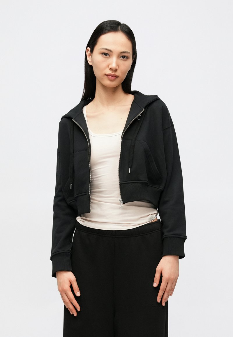 Camisola preta cropped com fecho de correr, bolsos canguru e textura suave, combinada com uma camisola interior leve e calças de fato de treino pretas.