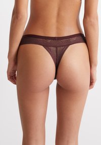 Tanga in pizzo borgogna caratterizzato da motivi geometrici, una vita morbida e un design posteriore minimal con dettagli trasparenti.
