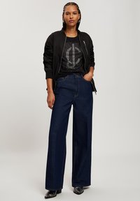 Czarna kurtka bomberka, pokryta cekinami czarna koszulka, szerokie ciemnoniebieskie dżinsy oraz błyszczące czarne buty. Model stoi z rękami w kieszeniach.