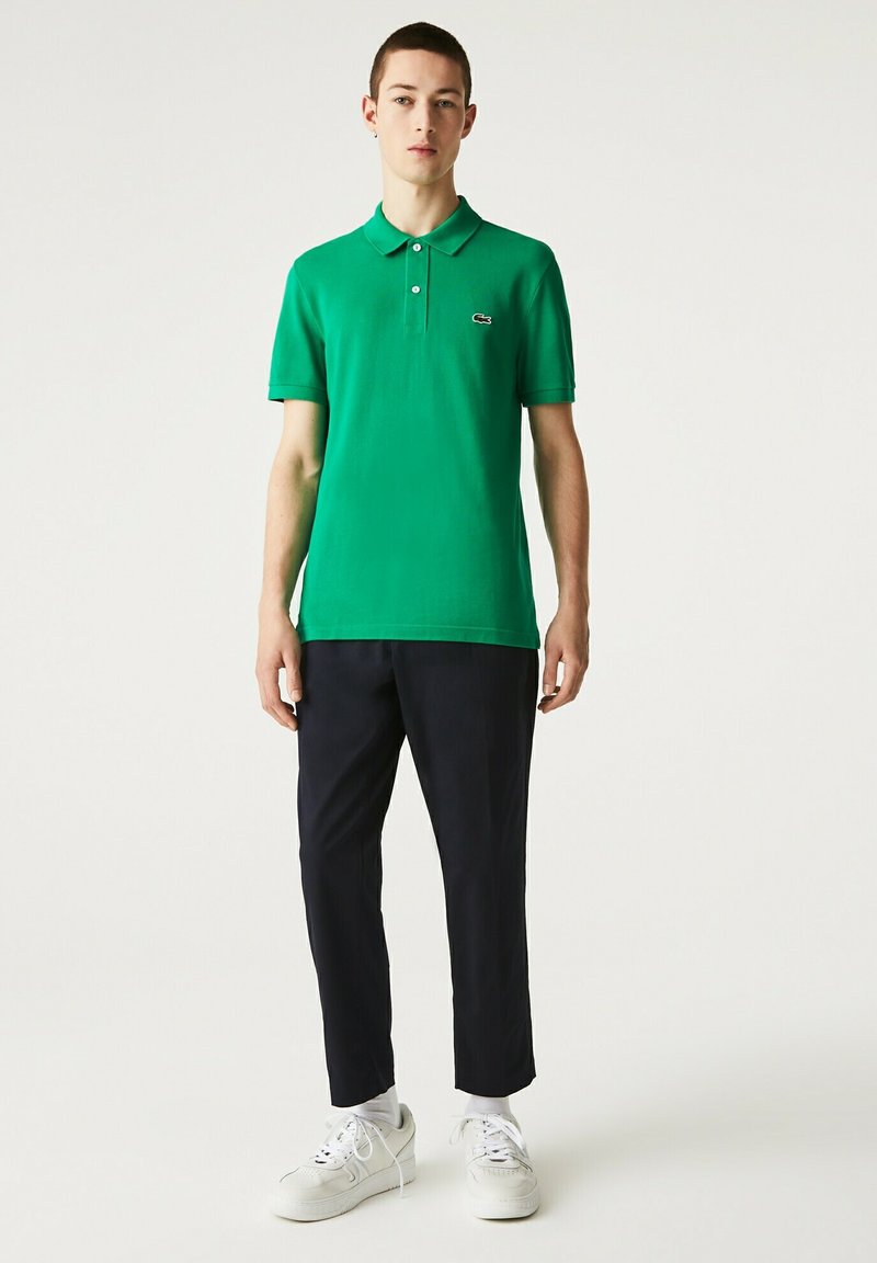 Grön polo skjorta med krage, korta ärmar och broderad logotyp. Bärs med marinblå byxor och vita sneakers, står mot en neutral bakgrund.