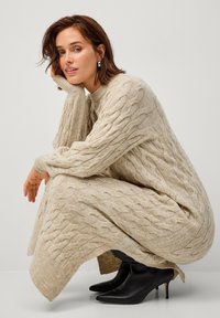 Beiges Strickkleid mit langen Ärmeln, strukturiertem Muster, Seitenschlitzen, kombiniert mit schwarzen spitzen Absatzstiefeln.