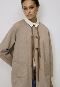 Beige oversized jas met een witte kraag, voorzien van een geknoopt voorpaneel met franjes en twee voorzakken. Textuur van een visgraatpatroon.