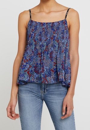 Femme portant un haut sans manches bleu à motifs floraux avec de fines bretelles noires, associé à un jean bleu clair, debout devant un fond uni.