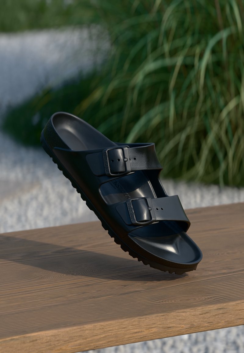 Sandale noire à double sangle avec boucles reposant sur une surface en bois, à côté de verdure et d'un fond de gravier blanc.
