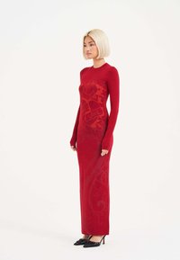 Vestido rojo de manga larga con silueta ajustada, diseño gráfico floral en un rojo más oscuro y cuello alto. Combinado con tacones negros de puntas.