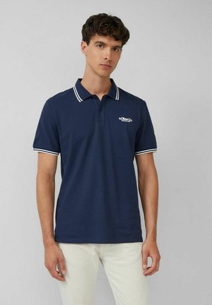 Junger Mann, der ein marineblaues Polohemd mit weiß gestreiftem Kragen und Ärmelbündchen trägt, kombiniert mit hellbeigen Hosen, steht vor grauem Hintergrund.