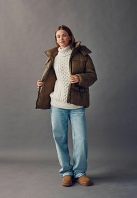 Puffet olivengrøn jakke med hætte over en struktureret hvid striktrøje, parret med lyseblå jeans og brune slip-on sko.