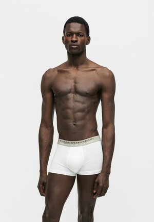 Izmos férfi sötét bőrrel, fehér Emporio Armani boxeralsót viselve, sima világos háttér előtt áll.