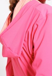 Prenda con capucha rosa de textura suave, que presenta un color de capucha interior en contraste y un diseño de corte relajado. Detalles de costura visibles.
