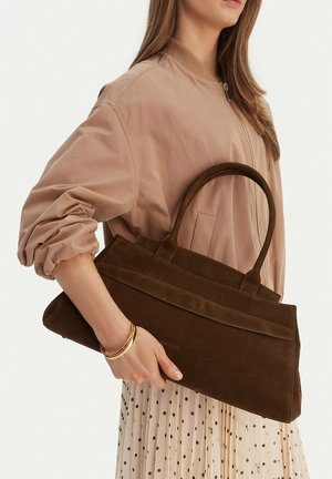 Žena v béžovém saku a sukni s puntíky drží velkou tmavě hnědou semišovou kabelku s dvojitými uchy a na zápěstí má zlaté náramky.