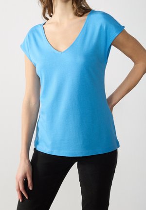 Femme portant un t-shirt bleu à manches courtes et col en V, et un pantalon noir, debout avec une main sur la hanche et l'autre détendue.