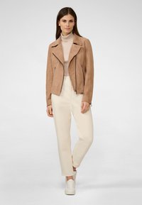 Beige mockaskinnmotorcykeljacka med asymmetrisk dragkedja, parat med krämfärgade byxor och vita sneakers. Slät textur och åtsittande design.