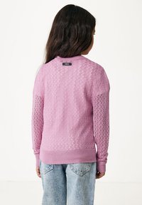 Ljust rosa stickad tröja med ett mönstrat mönster, med långa ärmar och ribbad nederkant. Lappen på ryggen säger "MEXX." Kombinerad med ljusblå jeans.