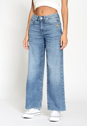 Femme portant un jean large bleu clair, un top court en maille blanche et des baskets compensées blanches sur un fond uni.