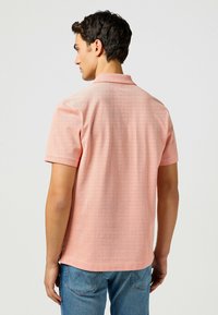 Polo a maniche corte di colore rosa chiaro con un motivo a righe testurizzato. Presenta un colletto e spacchi ai lati dell'orlo per favorire il movimento.
