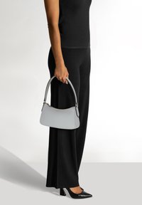 Borsa in pelle grigia di forma arrotondata, dotata di una maniglia corta e dettagli hardware sottili. Indossata con un abbigliamento e scarpe nere.