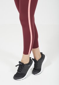 Leggings sportivi bordeaux con una striscia laterale rosa e la scritta "ENDURANCE". Abbinati a scarpe da atletica nere con suole bianche.