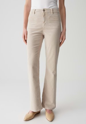 Beige Corduroy-Hose mit geradem Bein, die vordere Taschen, einen Knopfverschluss und subtile Nähedetails aufweist. Getragen mit hellen Schuhen.