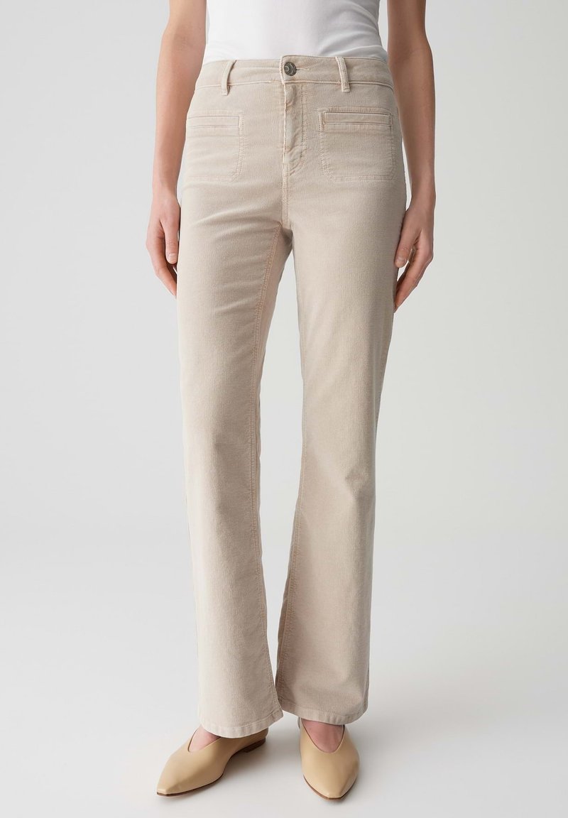 Beige Corduroy-Hose mit geradem Bein, die vordere Taschen, einen Knopfverschluss und subtile Nähedetails aufweist. Getragen mit hellen Schuhen.