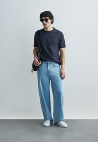 T-shirt às riscas pretas, jeans de perna larga azul claro, ténis brancos; complementado com óculos de sol pretos e uma bolsa transversal.