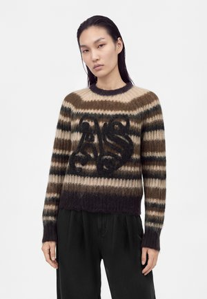 Personne aux cheveux raides et foncés porte un pull duveteux rayé marron et beige avec de grandes lettres noires stylisées à l'avant, associé à un pantalon noir ample.
